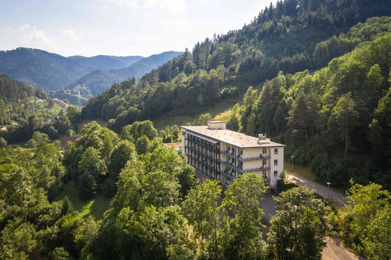 Panoramablick auf die Oberberg Fachklinik Schwarzwald: gro&szlig;es wei&szlig;es Klinikgeb&auml;ude in gr&uuml;ner H&uuml;gellandschaft mit B&auml;umen und blauem Himmel