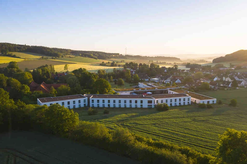 Au&szlig;enaufnahme der Oberberg Fachklinik Weserbergland: Geb&auml;ude mit wei&szlig;er Fassade und roten Akzenten, umgeben von B&auml;umen, Rasenfl&auml;che und Weg im Vordergrund