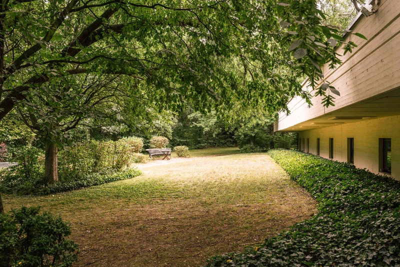 Gr&uuml;ner Gartenbereich mit gro&szlig;en B&auml;umen, Rasenfl&auml;che und einer Holzbank, rechts ein Geb&auml;udefl&uuml;gel der Oberberg Somnia Fachklinik K&ouml;ln-H&uuml;rth im Hintergrund